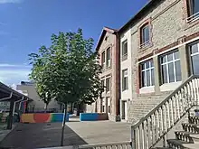 On voit au premier plan, derrière des barrières, une cour d'école avec un petit arbre et un muret coloré un peu plus loin. À gauche on devine un préau. Au second plan se dresse le bâtiment qui abrite l'école en elle-même ainsi que la mairie du village au dernier étage. L'ensemble est plutôt gris mais de larges fenêtres font en sorte que le tout ne semble pas trop triste.