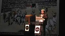 Photographie d'un homme parlant, debout derrière un pupitre.