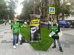 Photographie en couleurs de deux femmes portant des T-shirts verts et distribuant des tracts en ville.