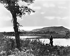 Photographie en noir et blanc d'une plaine une rivière qui y coule, un homme qui patrouille et une colline en fond.