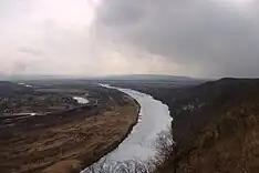 Photographie d'une rivière dans un méandre causé par un relief, avec des nuages gris dans le ciel.