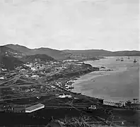 Photographie en noir et blanc d'une petite ville installée sur des monts au bord d'une baie où se trouve des bateaux.