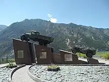 Photographie du monument de la légende des années 1930. Quatre blocs portent en tout deux véhicules, avec les blocs qui montent en hauteur. Le premier véhicule est plus imposant que le second.