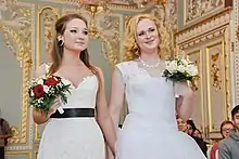 Photographie de deux femmes blondes se tenant la main, toutes deux en robe de mariée blanche
