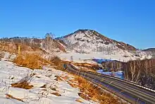 Photographie d'un espace enneigé avec des arbres ayant pour certains encore leurs couleurs d'automne, où la route circule. En arrière-plan se trouve une montagne de faible altitude.
