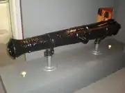 Photograph of the body of a black muzzle-loading cannon propped by two braces rest on a rectangular gray stand with two embedded little round lamps.