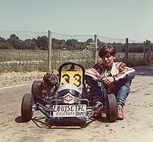 Photo plan large d'un kart noir flanqué du numéro 33 sur fond jaune. À côté, un jeune homme assis en combinaison de course, assis.