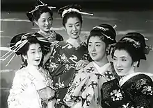 Cinq femmes souriantes prennent la pose, elles sont vêtues kimonos et leurs cheveux sont ornés de kanzashi (ornements traditionnels japonais) (image en noir et blanc).