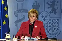 Photographie en couleur d'une femme vêtue d'une veste rouge. Elle est assise derrière un bureau. Devant elle est posé un microphone. Une cruche d'eau et un verre sont posés à sa droite. Sur sa droite est placé un drapeau bleu comportant des étoiles jaunes.