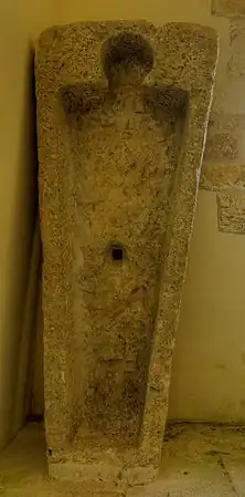 Photo couleur d'un bloc de pierre placé verticalement. Il est creusé pour accueillir une dépouille funéraire. Une loge plus petite est découpée pour recevoir le cou et la tête.