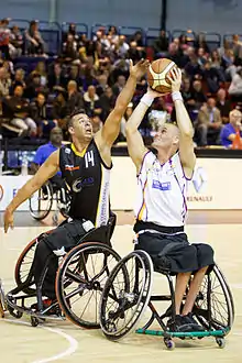Joueur de basket fauteuil tirant au panier sous la pression d'un défenseur au poste bas.
