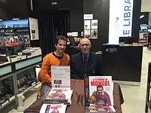 Un homme en costume cravate est assis devant une table présentant des livres, accompagné d'un jeune homme en tenue plus décontractée.