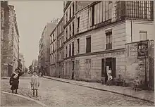 photographie sépia de la rue ; au premier plan se trouve une enseigne "crèmerie de la butte Montmartre" devant laquelle une femme est assise. Plusieurs enfants sont dehors, et des adultes se tiennent sur le pas des portes