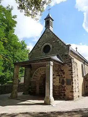 Photographie de la façade d'une chapelle romane. Entrée surmontée d'une voute en pierres alternativement claires et sombres, protégée par un auvent porté par deux colonnes carrées. Un œil de bœuf au-dessus de l'auvent ; un petit clocheton en faîte.