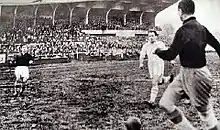 Joueur en noir avec le ballon proche de lui, avec un adversaire à quelques mètres, et un coéquipier plus loin.