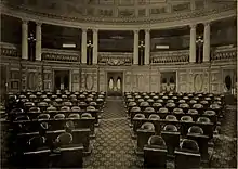 Illustration d'une assemblée avec un parterre de chaises devant une façade avec des piliers.
