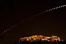 La Lune traverse l'image, passant d'une couleur grise à rouge puis grise. L'acropole est visible éclairée en bas.