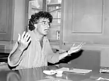 Photo noir et blanc d'un homme dans une salle en train de parler devant une table.