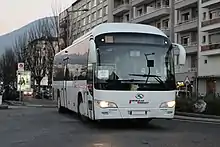 Photographie en couleurs d’un autocar blanc roulant dans une rue urbaine, devant un immeuble résidentiel.