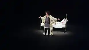 Une actrice sur scène, bras ouverts, illuminée par un projecteur, avec un lit derrière elle.