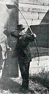 Photo d'un homme en costume et haut de forme s'apprêtant à décocher une flèche avec un arc.
