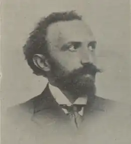 Photographie en noir et blanc d'un portrait en tête d'un homme barbu et moustachu. Sa tête est tournée vers sa gauche.