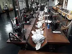Photographie en vue rapprochée d'une rangée de postes de travail des ouvrières chemisières du XIXe siècle reconstitués au musée. Un unique meuble long en bois accueille deux rangées de places qui se font face, chacune pourvue d'une machine à coudre. Des séparateurs en métal arborant la marque Singer séparent les places. Au centre, une rigole creuse dans le bois accueille le linge. Chaque place dispose d'un tabouret.