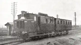 Photographie en noir et blanc de l'autorail de forme parallélépipèdique vue de trois quart face, côté gauche visible. Il est accompagné de sa remorque.