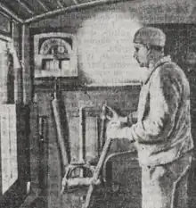 Photographie en noir et blanc de la cabine d'un autorail. Le conducteur est debout, porte un petit bonnet et tient dans sa main un levier. Sur la paroi de la cabine, un appareil enregistreur de vitesse est présent.