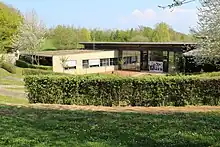 Bâtiment dont les couleurs font que ce dernier se fond dans le paysage dominé par des arbres verts et de l'herbe.