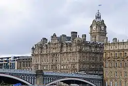 Un grand bâtiment rectangulaire de pierre grise comportant une multitude de fenêtres et une grande tour carrée en arrière plan comportant une horloge. Un pont est visible au premier plan et traverse un espace entre ce bâtiment est un autre de même style