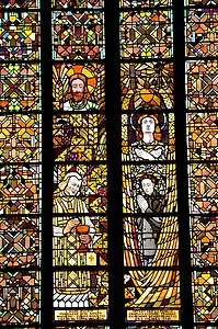 Elément de vitrail. Quatre personnages sont présents. À gauche, un homme barbu au-dessus d'un vieux prêtre. À droite, un jeune homme avec une longue chevelure au-dessus d'une jeune femme en armure.