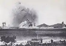 Photographie en noir et blanc d'une explosion sur la rive d'une baie vue depuis l'autre côté de la même baie.