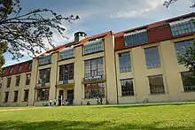 Photographie en couleur d’un bâtiment sur trois niveaux et pourvu de larges fenêtres. La façade est beige, le toit est rouge. Le premier plan montre un espace vert.