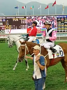Photo d'un jockey habillé en rose et noir sur un cheval alezan que tient un groom sur un hippodrome.
