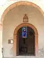 Le porche et l'entrée de l'église. Au-dessus de l'entrée est inscrite la date de 1872. Le vitrail visible dès l'entrée de l'église représente le Sacré-Cœur de Jésus.
