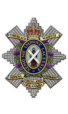 Image en couleur d'un insigne militaire composé d'une croix diamantée avec des listels d'or en haut et en bas ainsi qu'une couronne au naturel en haut. Au centre de l'insigne il y a un homme portant une robe brune tenant une croix blanche sur fond bleu entouré d'un anneau bleu foncé liséré d'or portant une inscription d'or entouré de chardon au naturel.