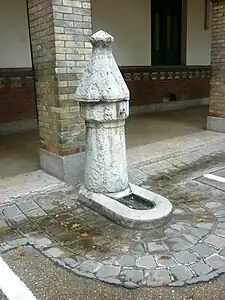 Fontaine à pompe : une borne en forme de pompe surmontant un puits déverse l'eau par un conduit dans une auge monolithique.