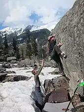 Un grimpeur sur un bloc en extérieur et une personne à côté du crash-pad qui fait une parade.