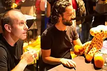 A man with short brown hair, sitting next to a man with curly black hair hugging a plush giraffe, both smiling at something to the right of the camera.