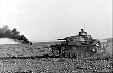 Panzer III avec un Kwk 38 sur le front africain passant devant un camion en feu (Afrique du nord, Avril 1941)
