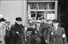 Des hommes se tiennent debout devant la vitrine d’une boutique et regardent dans la direction du photographe. Deux portent des brassards et des casquettes, et l’un d’entre eux brandit un bâton.