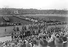 Photo noir et blanc d’un défilé de type militaire, avec des oriflammes, devant un public nombreux dans les tribunes du stade.