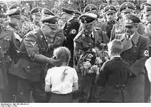 Photographie montrant deux enfants, une fille à gauche et un garçon à droite remettant un bouquet de fleurs à Heinrich Himmler au centre de la photographie ; le personnage de gauche est Wilhelm Koppe, celui de droite Fritz Bracht.