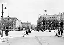 Photographie en noir et blanc d'une place traversée par des piétons et voitures, au fond des bâtiments.
