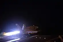 Photo de nuit d'un avion de chasse catapulté depuis un porte-avion. La scène est éclairée par la post-combustion des réacteurs, le reste du pont d'envol est plongé dans le noir.
