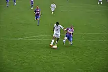 Joueur en train de tirer vers le but, un défenseur essayant de le contrer en le taclant par le côté.