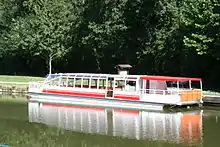 Photo en couleurs. Le bateau-promenade l'Oussance, vu de côté, est amarré sur la berge enherbée du port de Pont-aux-Moines, sur la commune de Mardié. Toute la partie avant est fermée et vitrée, permettant aux touristes de voir le paysage. L’arrière est constitué d’un pont ouvert recouvert d’une bâche rouge. Une large bande rouge tout le long du bateau, blanc par ailleurs, marque la ligne de flottaison.