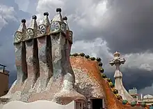 En quatre plans : groupe de cheminées polychromes, toit de la Casa Batlló, croix gaudienne, ciel d'orage.