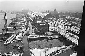 La gare centrale, vue depuis le nord-ouest, un jour de neige. Elle est bordée, à gauche par un terminal de bacs. Au premier plan, le Westertoegang (nl), court canal réunissant le lac IJ, à gauche, et le canal Singel, à droite. Entre les deux grandes verrières de la gare, il existe encore une partie à ciel ouvert
